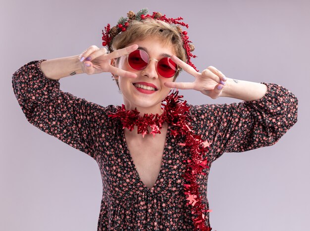 Joyful young pretty girl wearing christmas head wreath and tinsel garland around neck with glasses looking at camera showing v-sign symbols near eyes isolated on white background