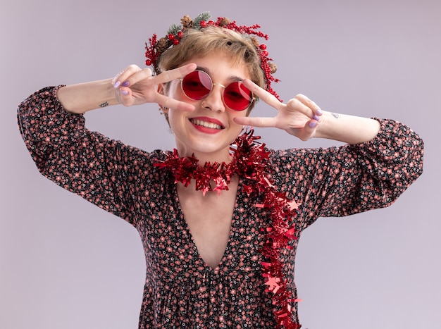 Joyful young pretty girl wearing christmas head wreath and tinsel garland around neck with glasses looking at camera showing v-sign symbols near eyes isolated on white background
