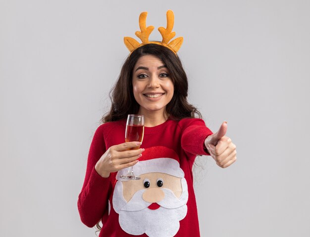 Joyful young pretty girl wearing reindeer antlers headband and santa claus sweater holding glass of champagne looking showing thumb up 