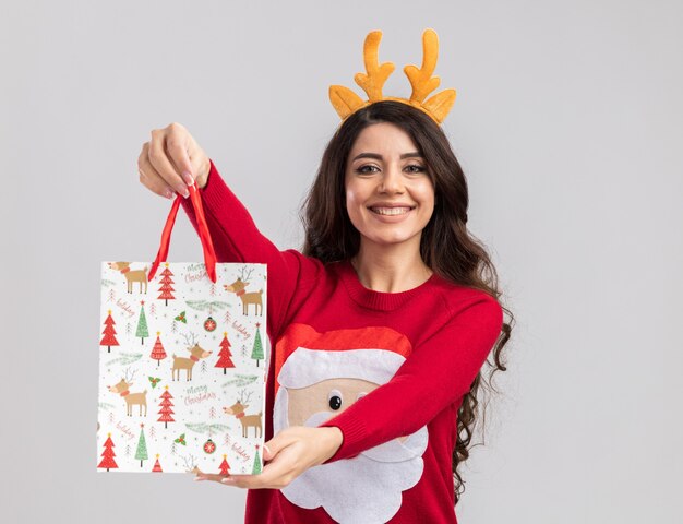 Joyful young pretty girl wearing reindeer antlers headband and santa claus sweater stretching out christmas gift bag towards camera looking 