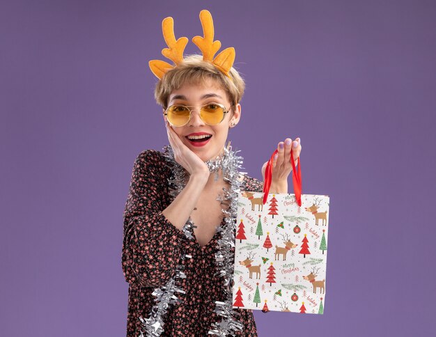 トナカイの角のカチューシャと見掛け倒しの花輪を首にかけ、クリスマス ギフト バッグを持った眼鏡をかけた、うれしそうな若い可愛い女の子が、コピー スペースを持つ紫の壁に顔を離さずに手をつないでいる