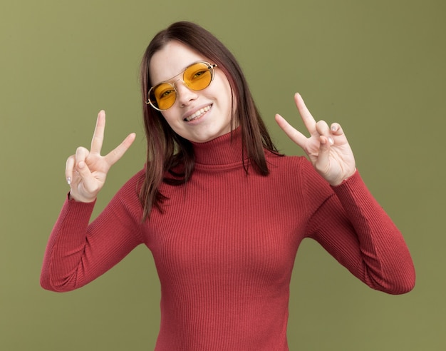 Joyful young pretty girl wearing sunglasses  doing peace sign isolated on olive green wall