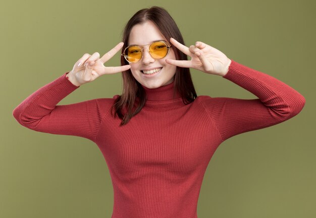 Joyful young pretty girl wearing sunglasses showing v-sign symbols near eyes 