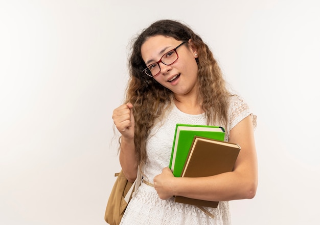 Joyful young pretty schoolgirl wearing glasses and back bag holding books clenching fist isolated on white