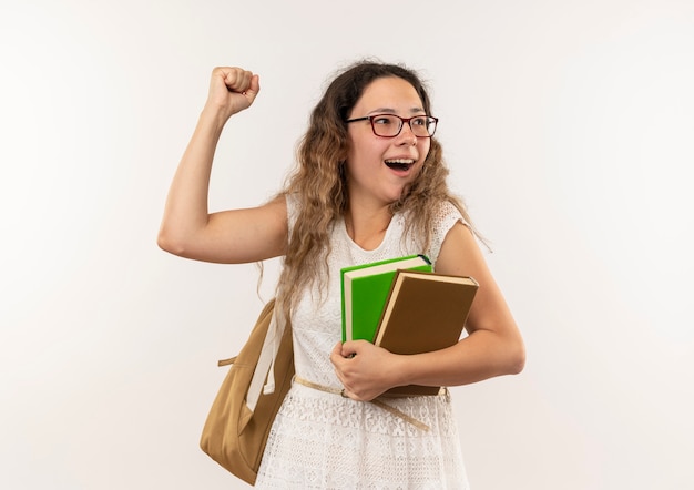 Радостная молодая симпатичная школьница в очках и задней сумке держит книги, глядя в сторону и поднимая кулак, изолированные на белом