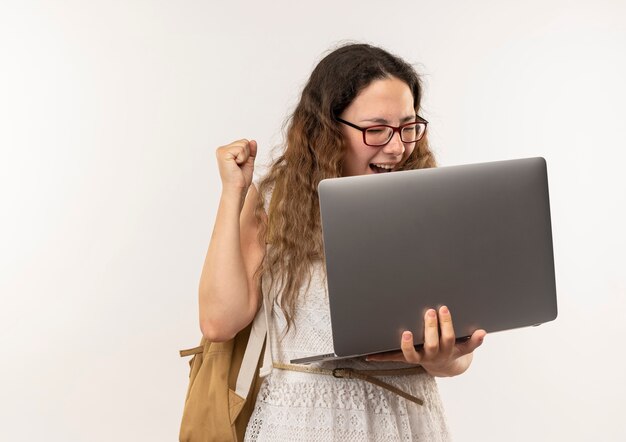 Joyful young pretty schoolgirl wearing glasses and back bag holding laptop raising fist isolated on