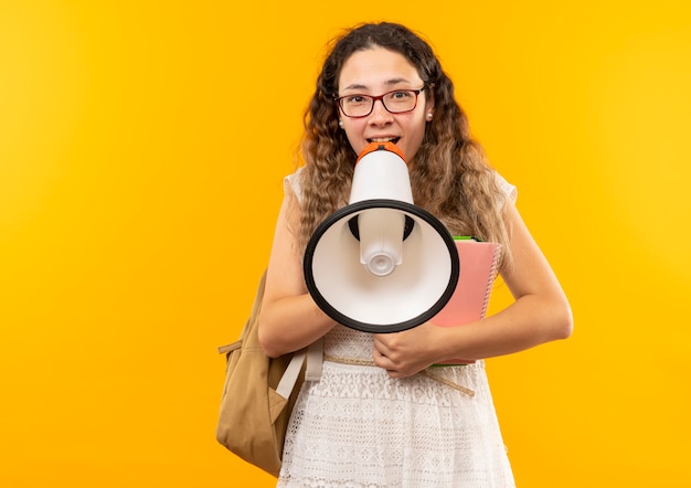 Free photo joyful young pretty schoolgirl wearing glasses and back bag holding note pad talking by speaker isolated on yellow