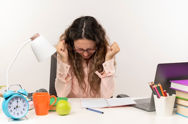 Foto gratuita gioiosa giovane studentessa graziosa con gli occhiali seduto alla scrivania con strumenti di scuola facendo i compiti stringendo i pugni con gli occhi chiusi isolati su bianco