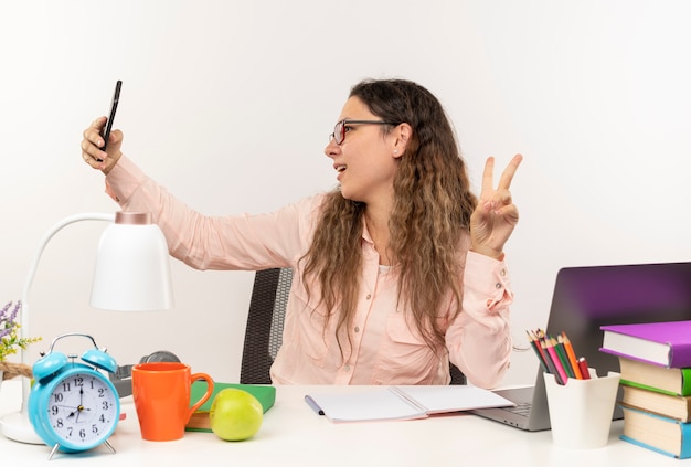 Foto gratuita gioiosa giovane studentessa graziosa con gli occhiali seduto alla scrivania con strumenti di scuola facendo i compiti facendo segno di pace prendendo selfie isolato su bianco