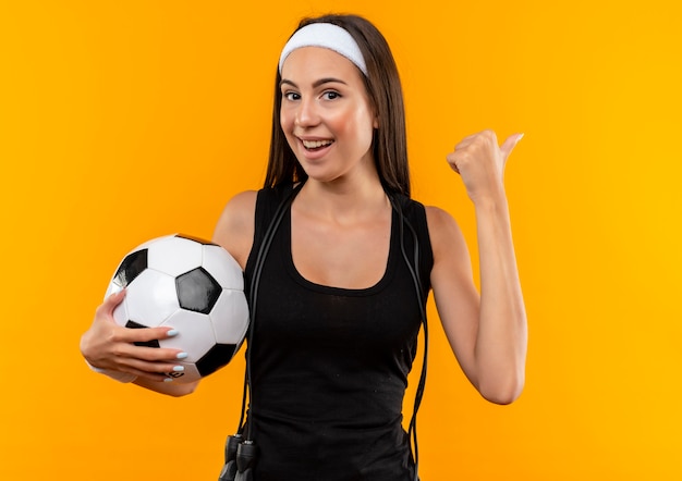 Бесплатное фото Радостная молодая симпатичная спортивная девушка с ободком и браслетом держит футбольный мяч и указывает в сторону со скакалкой на шее, изолированной на оранжевом пространстве