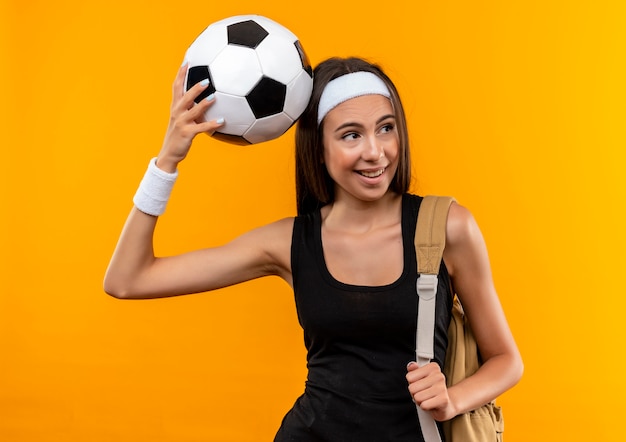 Радостная молодая симпатичная спортивная девушка с головной повязкой и браслетом и сумкой на спине кладет футбольный мяч на голову, глядя на сторону, изолированную на оранжевом пространстве
