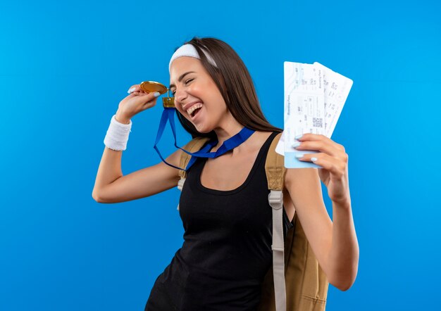 Joyful young pretty sporty girl wearing headband and wristband and back bag with medal around neck holding airplane tickets and medal with closed eyes isolated on blue space