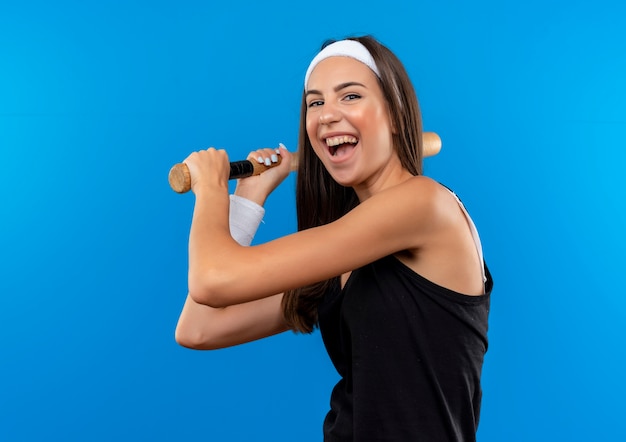 Joyful young pretty sporty girl wearing headband and wristband holding baseball bat isolated on blue space