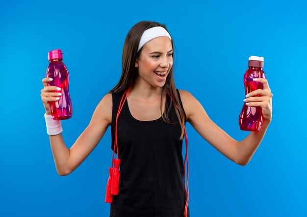 Joyful young pretty sporty girl wearing headband and wristband holding and looking at water bottles with jumping rope around her neck isolated on blue space