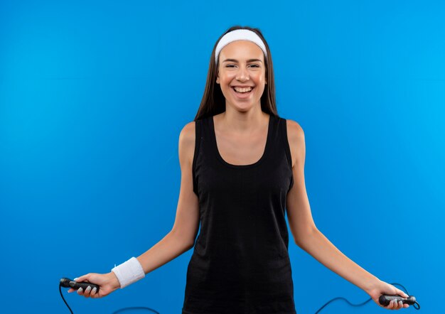 Joyful young pretty sporty girl wearing headband and wristband jumping rope isolated on blue space