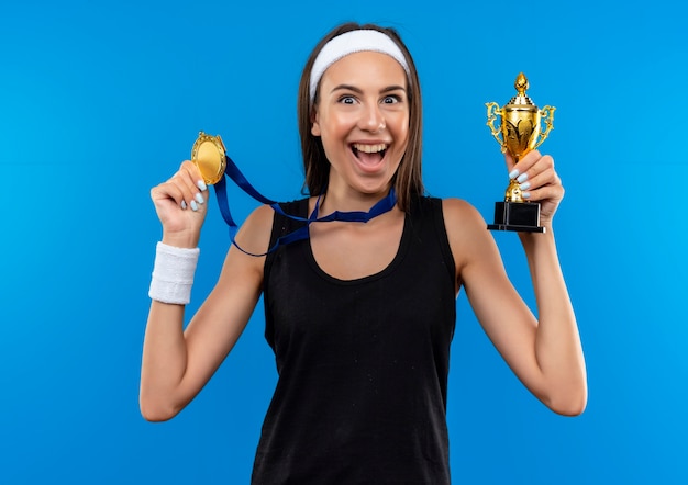 Joyful young pretty sporty girl wearing headband and wristband and medal around her neck holding winner cup and medal isolated on blue space