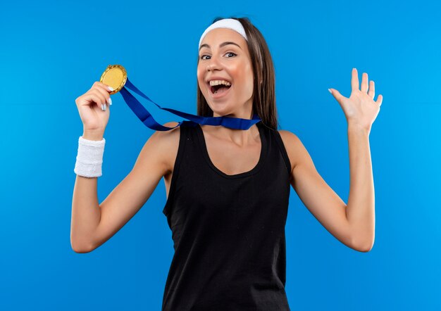 Радостная молодая симпатичная спортивная девушка с головной повязкой и браслетом и медалью на шее держит медаль и показывает пустую руку, изолированную на синем пространстве
