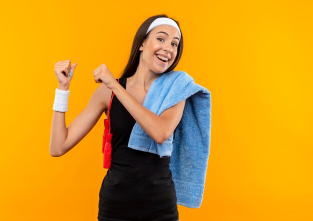 Joyful young pretty sporty girl wearing headband and wristband with towel and jumping rope on her shoulders pointing behind on orange space 