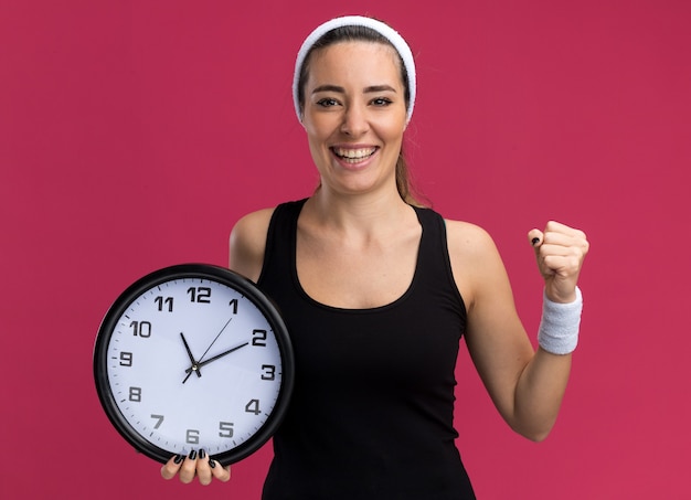 Joyful young pretty sporty girl wearing headband and wristbands holding clock  doing be strong gesture isolated on crimson wall