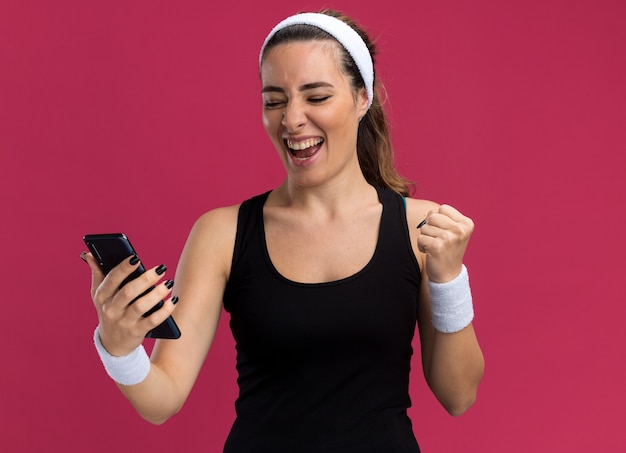 Радостная молодая симпатичная спортивная девушка с повязкой на голову и браслетами, держащая и смотрящая на мобильный телефон, делает жест `` да ''