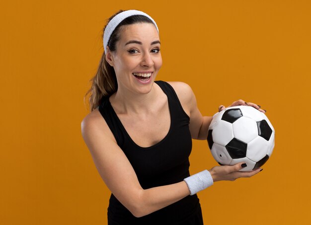 Joyful young pretty sporty girl wearing headband and wristbands holding soccer ball isolated on orange wall with copy space