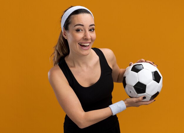 コピースペースでオレンジ色の壁に分離されたサッカーボールを保持しているヘッドバンドとリストバンドを身に着けているうれしそうな若いかなりスポーティーな女の子