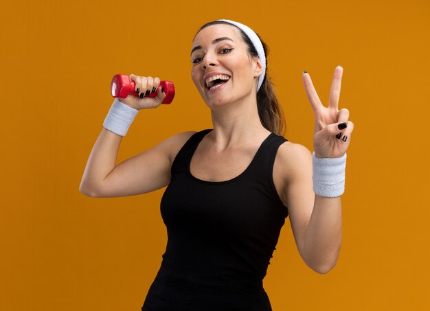 Joyful young pretty sporty woman wearing headband and wristbands holding dumbbell looking at front doing peace sign isolated on orange wall