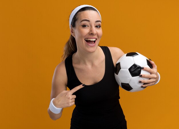 Joyful young pretty sporty woman wearing headband and wristbands holding soccer ball pointing at it looking at front isolated on orange wall with copy space