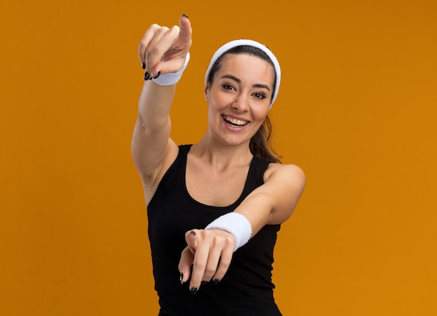 Foto gratuita gioiosa giovane donna piuttosto sportiva che indossa fascia e braccialetti che guardano davanti facendo un gesto isolato sulla parete arancione con spazio per le copie