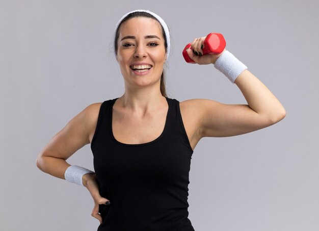 Joyful young pretty sporty woman wearing headband and wristbands looking at front keeping hand on waist raising dumbbell isolated on white wall