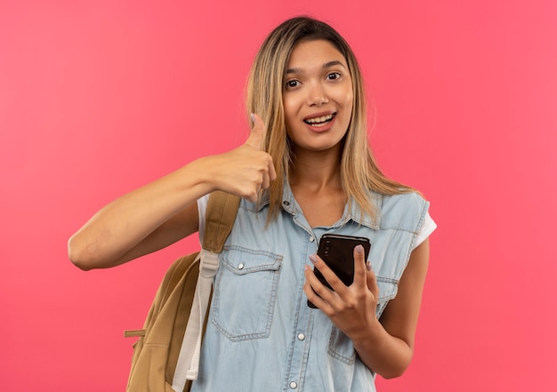 Радостная молодая симпатичная студентка в сумке на спине, держащая мобильный телефон и показывающая большой палец вверх изолирована на розовом