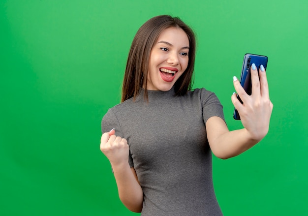 Joyful young pretty woman holding and looking at mobile phone and clenching fist isolated on green background with copy space