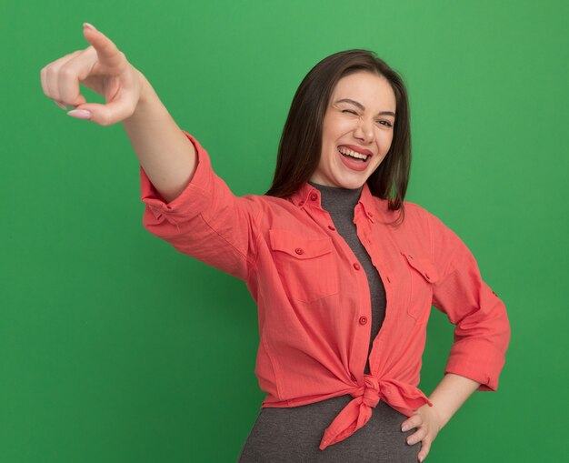 Joyful young pretty woman keeping hand on waist looking straight pointing at side winking isolated on green wall