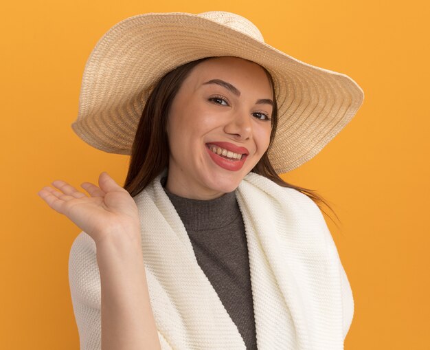 Joyful young pretty woman wearing beach hat  showing empty hand isolated on orange wall