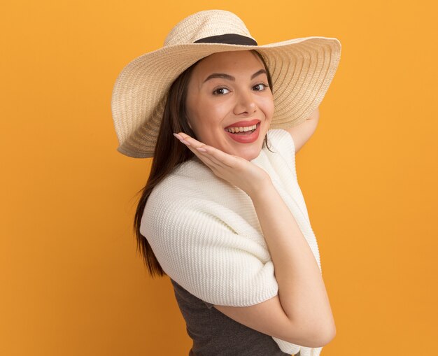 Joyful young pretty woman wearing beach hat standing in profile view  touching chin isolated on orange wall