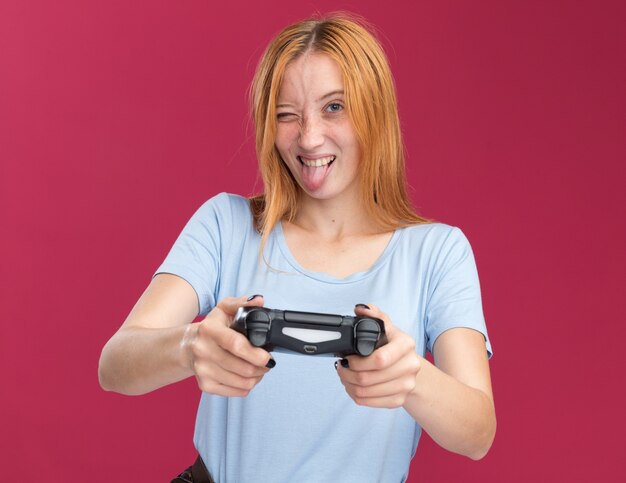 そばかすのあるうれしそうな若い赤毛の生姜の女の子は舌を突き出し、ピンクのゲームコントローラーを保持します