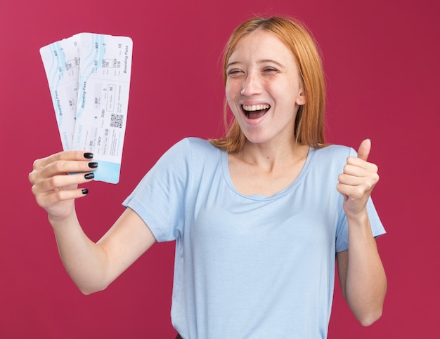 joyful young redhead ginger girl with freckles thumbing up and holding air tickets isolated on pink wall with copy space