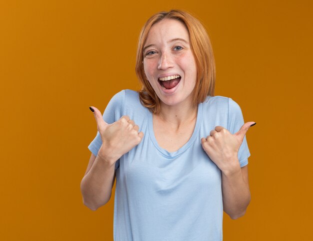 Joyful young redhead ginger girl with freckles thumbing up with two hands on orange