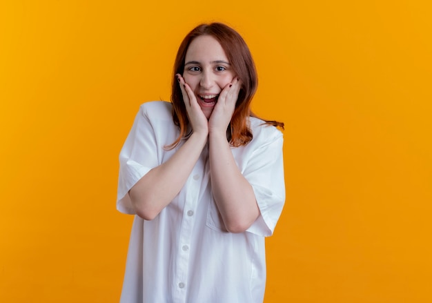 Joyful young redhead girl putting hands on cheek isolated on yellow background