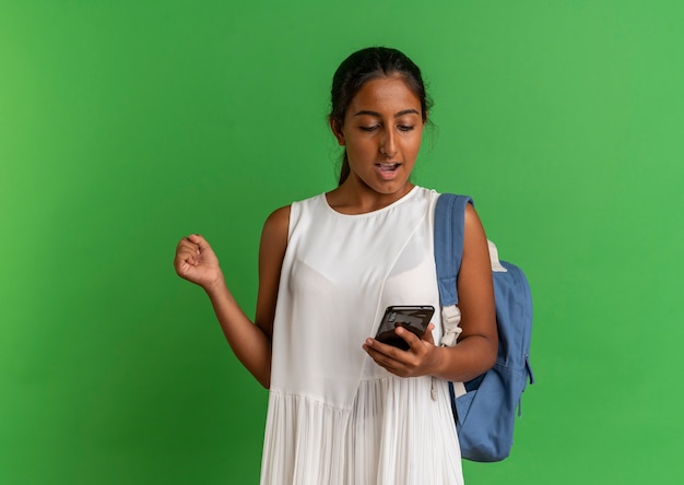 Joyful young schoolgirl wearing backpack holding and looking at phone and showing yes gesture 