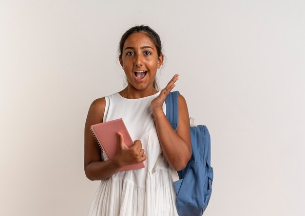 Joyful young schoolgirl wearing backpack holding notebook and raising hand 