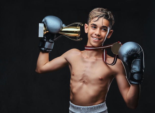 Joyful young shirtless boxer champion wearing gloves holds a winner's cup and the gold medal. Isolated on the dark background.