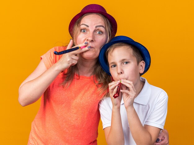 радостный молодой славянский мальчик в синей партийной шляпе и его мать в фиолетовой партийной шляпе дует партийный свисток на оранжевой стене с копией пространства