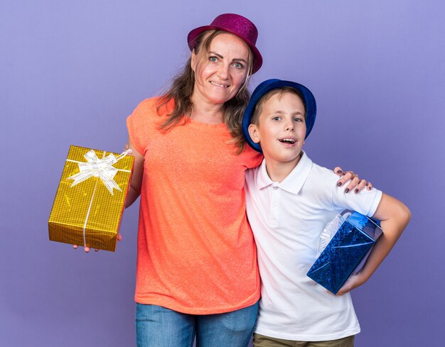 コピースペースと紫色の壁に分離された紫色のパーティーハットを身に着けている彼の母親とギフトボックスを保持している青いパーティーハットを持つ楽しい若いスラブ少年