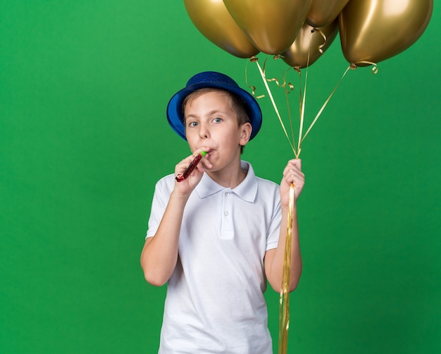 joyful young slavic boy with blue party hat holding helium balloons and blowing party whistle isolated on green wall with copy space