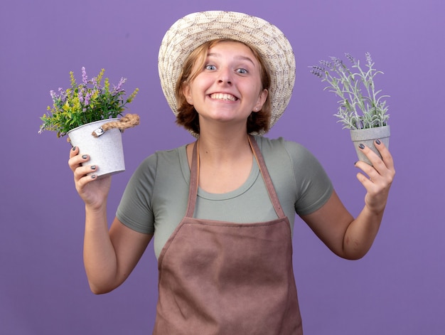 Free photo joyful young slavic female gardener wearing gardening hat holding flowers in flowerpots on purple