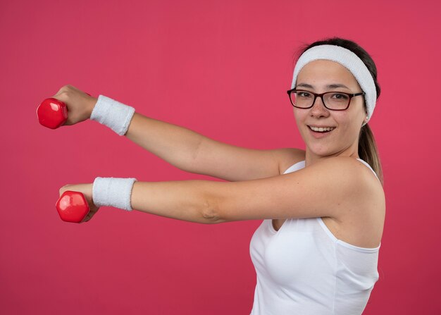 Радостная молодая спортивная девушка в оптических очках с повязкой на голову и браслетами стоит боком, держа гантели