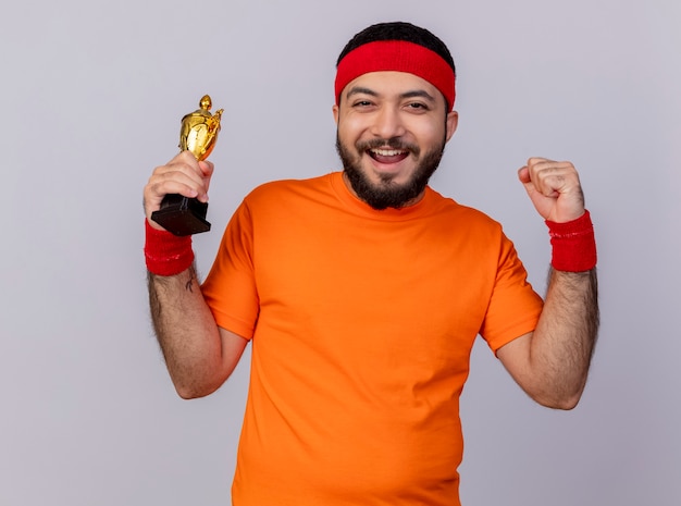 Joyful young sporty man wearing headband and wristband holding winner cup showing strong gesture isolated on white background