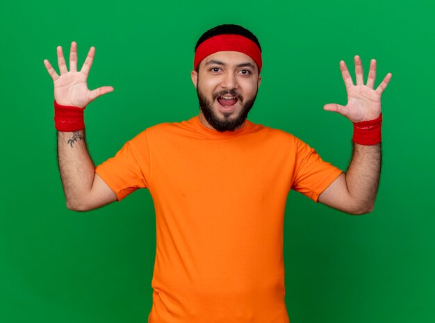 Free photo joyful young sporty man wearing headband and wristband raising hands isolated on green background