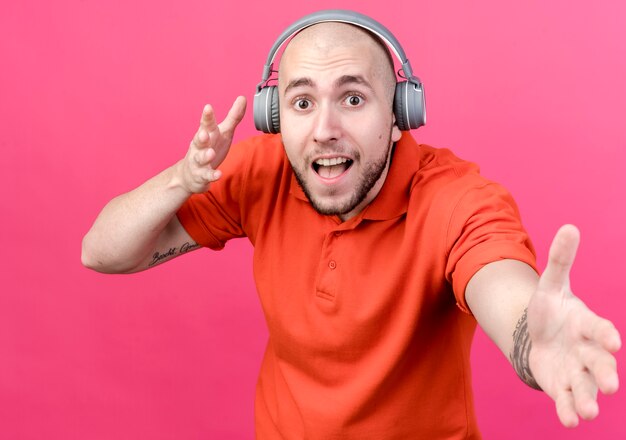 Joyful young sporty man wearing headphones and holding out hand  isolated on pink  wall